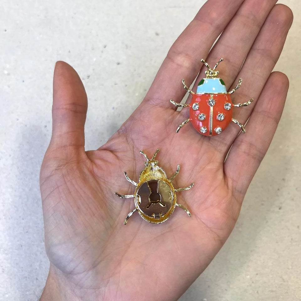 Dos cubre botones de mariquita en la palma de una mano: uno esmaltado en coral con cristales y otro mostrando el broche dorado por detrás